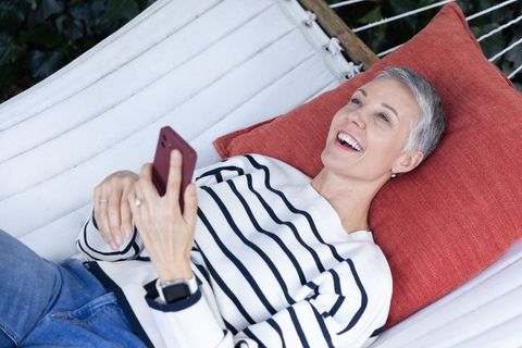 Senior woman relaxing in backyard hammock using smartphone, smiling in striped sweater