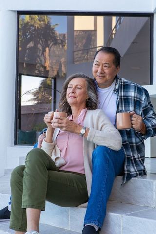 Diverse couple relaxing outside modern home with ceramic mugs