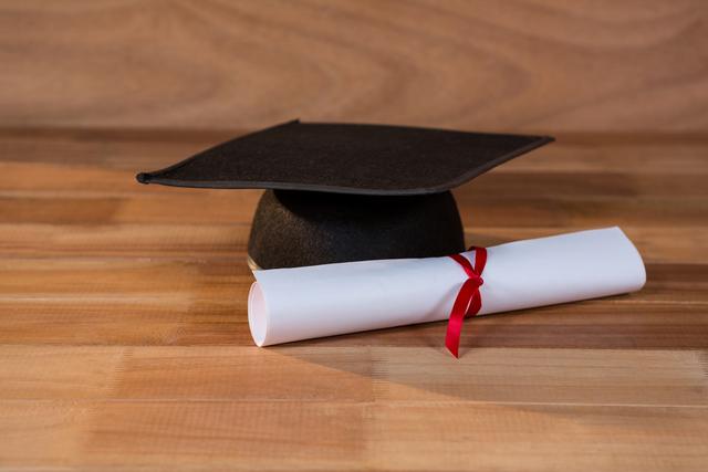 This image shows a graduation cap and a rolled diploma with a red ribbon on a wooden table. It can be used for educational materials, graduation announcements, academic achievements, and school or university promotions.