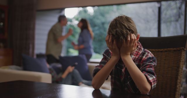 Young boy sitting at table covering his face with both hands while his parents argue in the background. Scene captures family conflict, emotional stress, and impact on children. Useful for topics around domestic issues, parenting challenges, counseling, and emotional well-being.