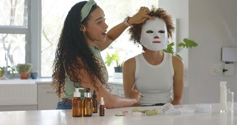 African american women adjusting led facial mask with skincare products in home kitchen