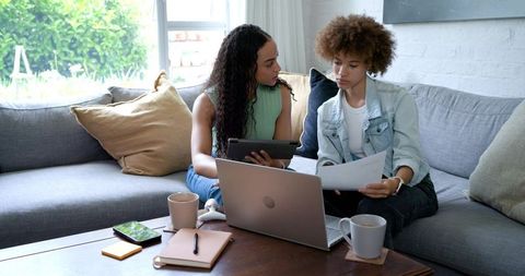 African american women collaborating on laptop and tablet at home, reviewing documents