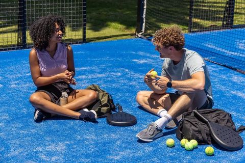 Mixed-race friends sitting and laughing on blue padel court with paddles and tennis balls
