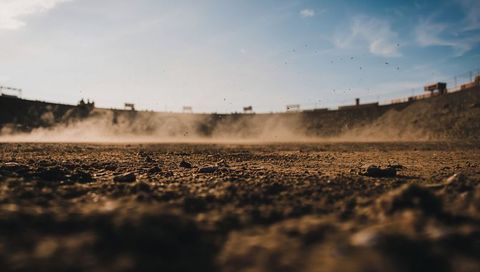 Low-Angle Dust Rising Over Textured Dry Arena Ground, Sunlit Perimeter Wall