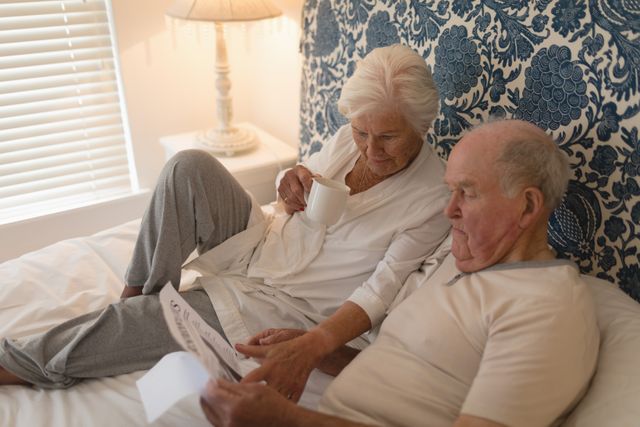 Senior couple enjoying a relaxed morning in bed, reading the newspaper and drinking coffee. Ideal for use in articles or advertisements about retirement, senior living, morning routines, and family bonding. Can also be used in healthcare and wellness contexts to depict a peaceful and healthy lifestyle for older adults.