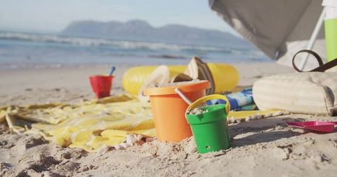 Colorful beach buckets resting on sunlit sand with towel, umbrella and playful summer toys