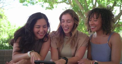 Multicultural friends laughing together and sharing smartphone on outdoor sofa