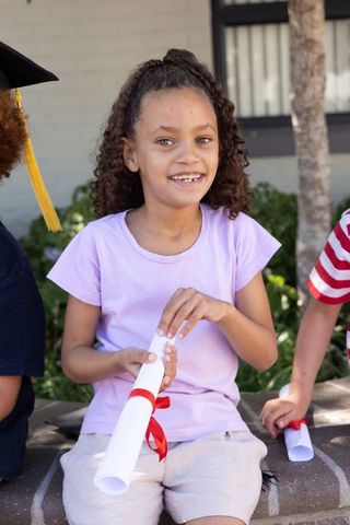 Smiling diverse girl holding diploma, celebrating elementary school graduation outdoors