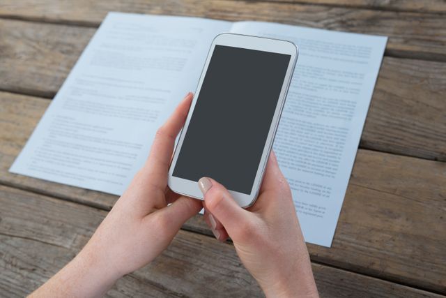 Businesswoman holding smartphone over documents on wooden table. Ideal for illustrating concepts of multitasking, modern business communication, remote work, and professional office environments.