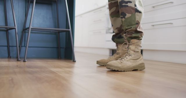 Large close-up view of military personnel wearing camouflage pants and army boots, standing on wooden flooring in a kitchen. This image can be used in articles about veteran life, stories about military personnel at home, or for material discussing military uniforms and lifestyle.