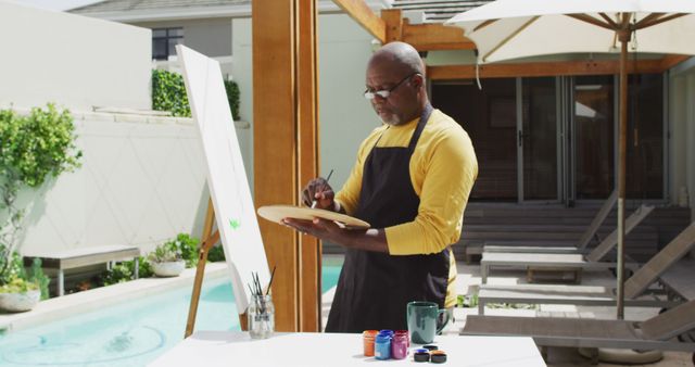 This image depicts a mature man engaged in painting on a canvas in a modern backyard next to a swimming pool. He is wearing a yellow shirt under an apron and is holding a palette, concentrating on his artwork. Ideal for themes related to creativity, hobbies, summertime, leisure activities, outdoor art sessions, or artistic inspiration.
