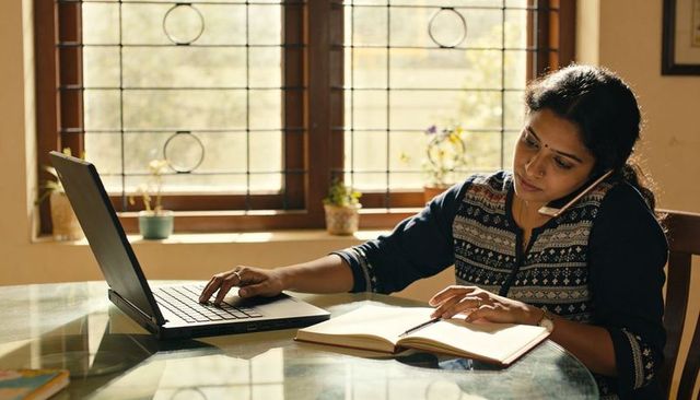 Typing woman in kurta holding phone and writing in notebook at sunlit home office