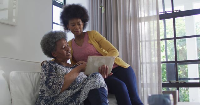 Older woman sitting on couch and young woman standing beside, both looking at tablet. Suitable for concepts of family bonding, technology use in day-to-day life, and intergenerational relationships. Ideal for articles, blog posts, and advertisements about technology, family life, and elder care.