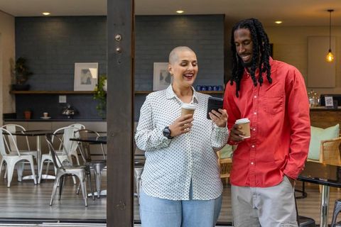 Smiling diverse couple enjoying takeaway coffee and checking smartphone in modern cafe