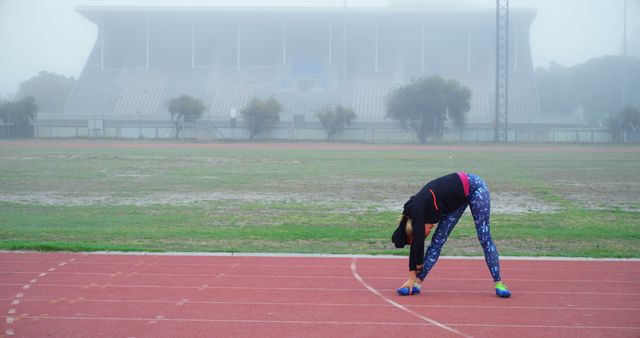 This image shows a woman in activewear stretching on a stadium track on a foggy day. The fog and deserted stadium create a moody atmosphere. This can be used for fitness magazines, advertising sportswear, promoting outdoor exercise, or illustrating dedication in athletic training.
