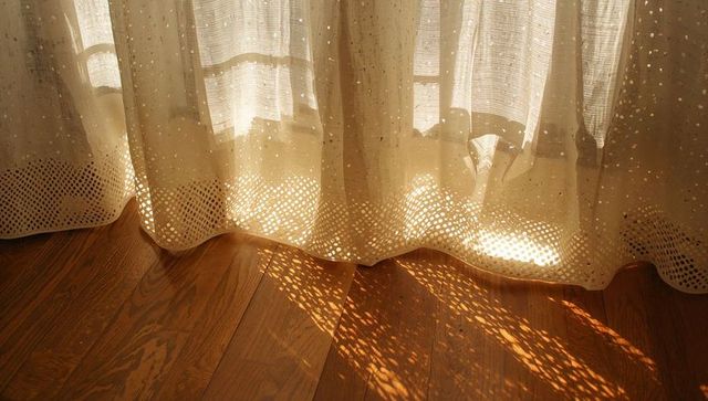 Sunlit sheer curtain casting dotted lace shadows on warm hardwood floor with scallop hem