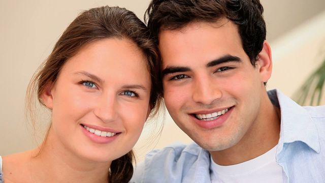 Cheerful young couple sitting together indoors, smiling at the camera. Perfect for use in personal care advertisements, relationship blogs, and social media campaigns promoting positivity and togetherness.