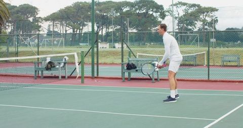 Male tennis player standing ready on outdoor court in white outfit holding racket