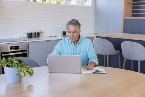 Senior man working remotely on laptop at round kitchen table in modern sunlit home office