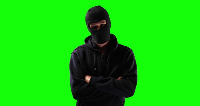 Image shows masked person wearing black hoodie with crossed arms standing against green screen background. Useful for themes of crime, anonymity, mystery, and security. Green screen makes it easy to edit and customize the background for various digital content purposes.