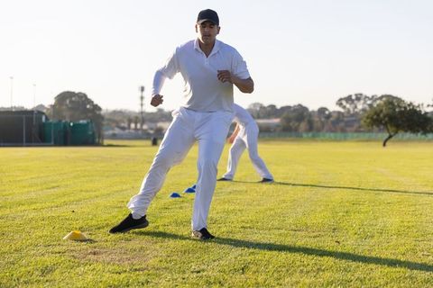 Cricket player performing lateral agility drill on sunlit field wearing white kit and cap