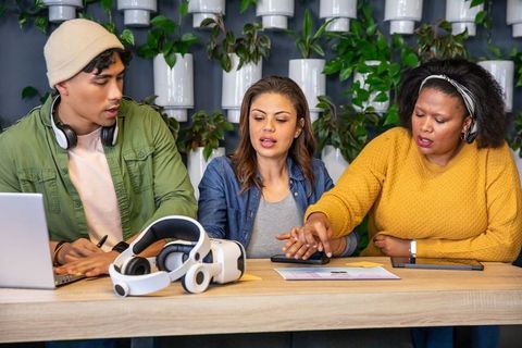 Multicultural team collaborating with vr headset, laptop and tablet in green workspace