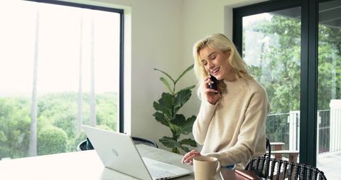 Remote worker smiling while talking on phone at bright home desk with laptop and coffee