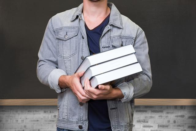 Midsection of male student holding a stack of books while standing in front of a blackboard. He is wearing a denim jacket and a dark shirt. Ideal for education-related websites, blogs about learning and teaching, college and school promotions, and visual content for academic content.
