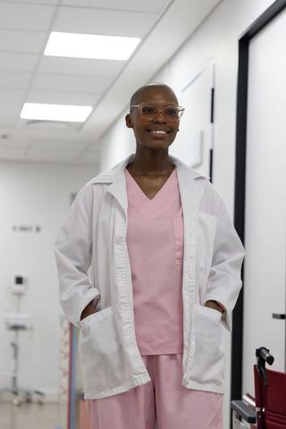 Smiling black nurse in pink scrubs and white lab coat standing in bright clinic hallway