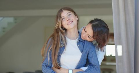 Same-sex couple embracing by window wearing denim jacket and smartwatch, intimate moment