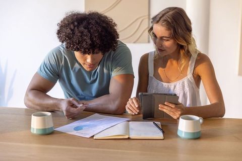 Young diverse couple reviewing budget at home using tablet, receipts and teal mugs