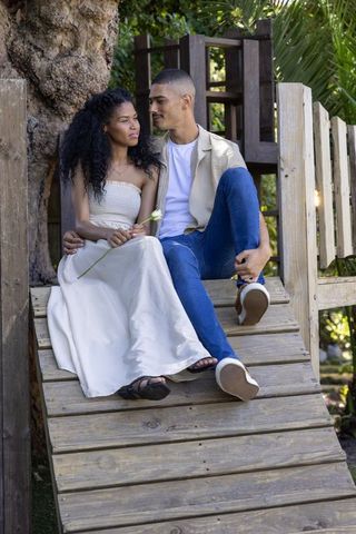 Romantic couple sitting on rustic wooden ramp holding white flower in tropical sunlight