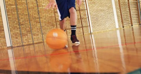 Teen dribbling basketball on polished hardwood gym floor with reflected sneakers