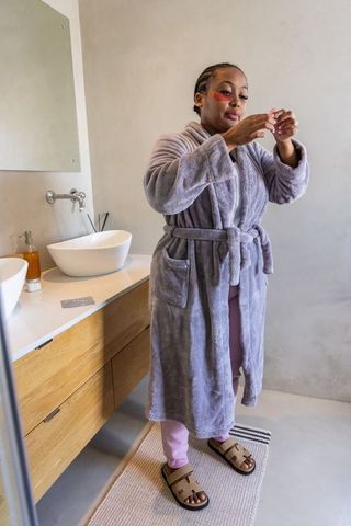 African american woman applying under-eye gel patches while holding skincare jar in cozy robe