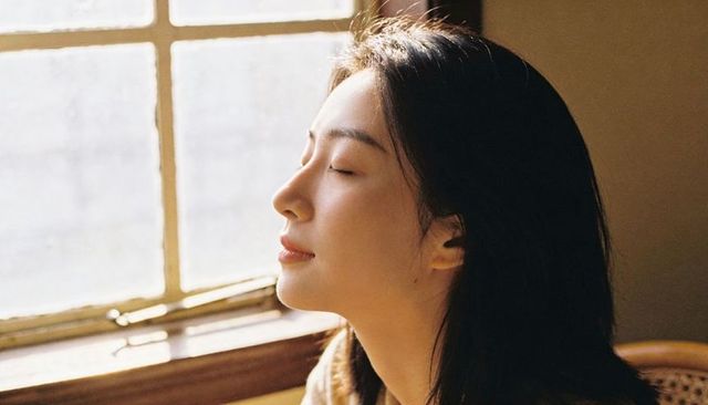 Sunlit woman basking by window in cozy beige sweater, serene minimalist interior