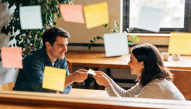 Colleagues exchanging sticky note during casual coworking brainstorming session by window