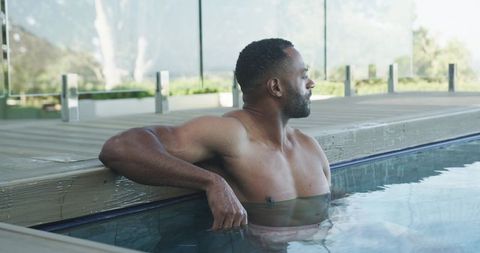 Mature man relaxing in sunlit outdoor pool, leaning on deck with glass railing