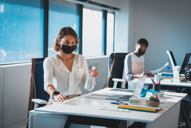 Two diverse businesswomen wearing face masks sitting at office desks, one disinfecting her workspace. independent creative business at a modern office during coronavirus covid 19 pandemic.