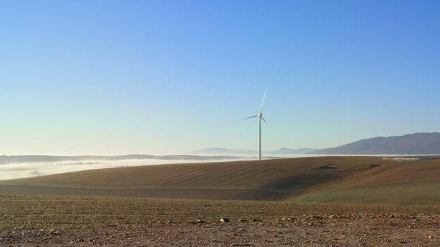 Wind turbine located on a farm with clear blue sky and gentle mist in background. Illustrates renewable and clean energy. Ideal for promoting sustainability, green energy initiatives, and rural landscapes.