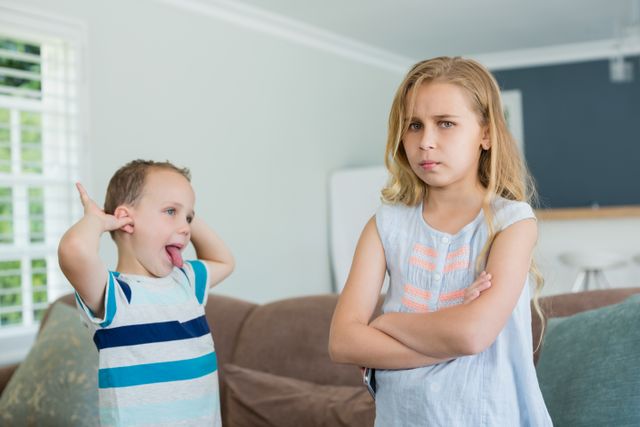 Brother teasing his sister while standing with arms crossed in living room at home