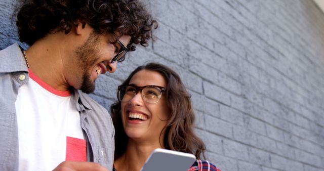 Happy diverse couple with smartphone, standing smiling by wall on sunny street. City break, summer, travel, vacations and lifestyle, friendship, technology, unaltered.