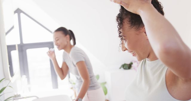 Two women are seen engaging in their morning routine in a bright bathroom. One is brushing her teeth while the other is styling her hair with a happy expression. The room is filled with natural light, contributing to a fresh and vibrant atmosphere. This image can be used to promote personal care products, lifestyle and health articles, morning routine tips, or women's health campaigns.