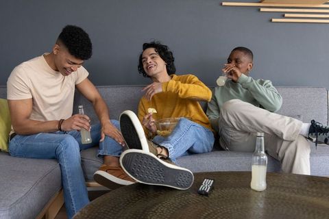 Young friends laughing and relaxing on sofa while drinking bottled beverages and snacking