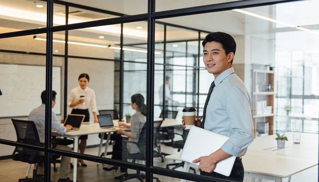 Confident executive holding coffee and laptop by glass meeting room in modern office