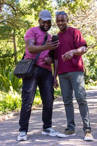 African men smiling while sharing smartphone on park path under dappled sunlight