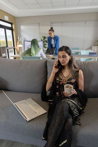 Woman working in modern coworking lounge calling while holding coffee with laptop nearby