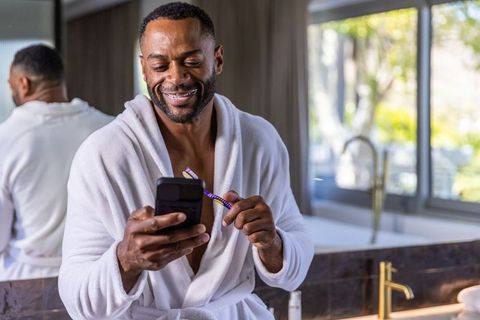 Smiling man wearing white bathrobe checking smartphone while holding toothbrush in bathroom