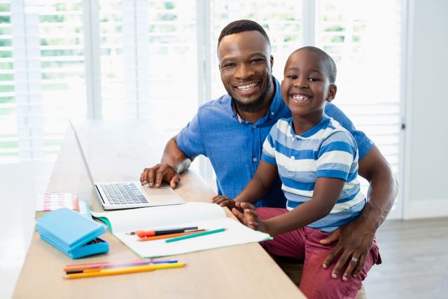 Father and son sitting at a table, father using a laptop while son is doing homework. Both are smiling and enjoying their time together. Ideal for use in family, education, and parenting contexts, showcasing positive family interactions and home learning environments.