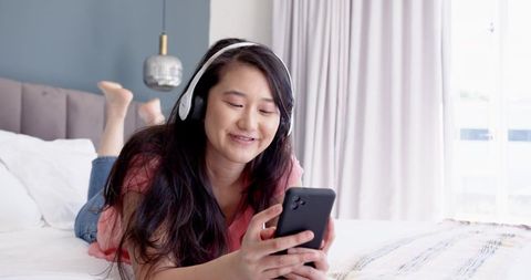 Asian woman relaxing in modern bedroom wearing headphones using smartphone for streaming