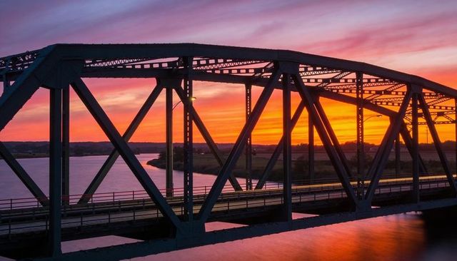 Steel truss bridge silhouetted at vibrant sunset over river with car light trail